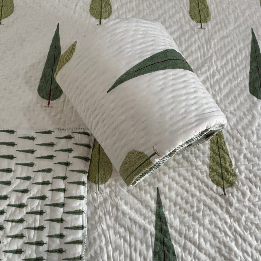 A close-up of a quilted white quilt with a green leaf pattern, showing a rolled section of the quilt and a folded corner with a different green pine-tree pattern.